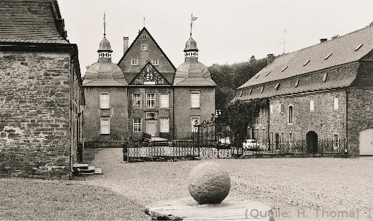 Foto: Von links nach rechts: Ein langgestrecktes Gebäude, rechts dahinter ein schloßähnliches Haus mit zwei Türmen. Rechts im Foto wieder ein langgestrecktes Gebäude. Der dadurch entstandene Hof ist vorne durch ein schmiedeeisernes Schmuckgitter begrenzt.