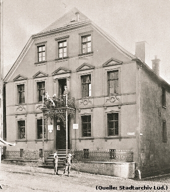 Foto: Ein altes Haus mit Treppenaufgang. Über der Haustür ein Balkon. Dort und vor dem Haus sind Personen zu erkennen.