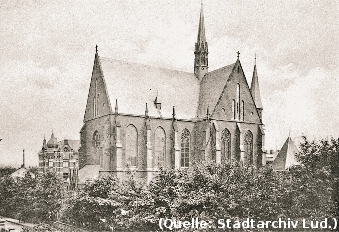 Foto: Eiune große Kirche erhebt sich hinter den Bäumen. Häuserdächer sind erkennbar.