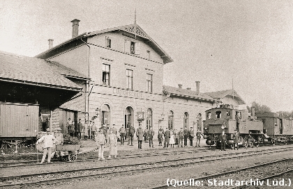 Foto: Ein Bahnhof. Davor Personen und ein Zug mit einer Dampflok.