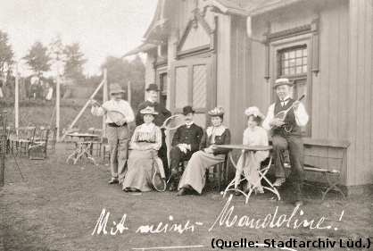 Foto: Um 1910: Eine Gesellschaft an einem Tennisplatz. Ein Mann spielt Mandoline auf seinem Tennisschläger.
