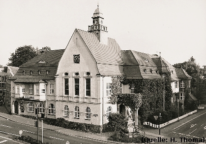 Foto: Ein Eckhaus an einer Strassenkreuzung, davor ein Brunnen.