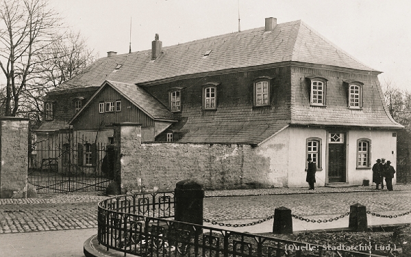 Foto: Ein kleines, zweistöckiges Haus mit umgebender Mauer. Davor einige Personen.