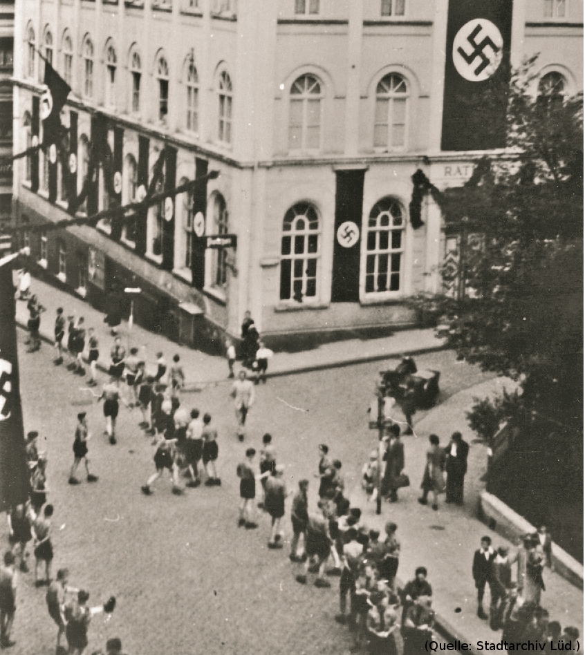 Foto: Ein Haus an einer Strasse. Das Haus ist behängt mit Hakenkreuzfahnen. Auf der Strasse Menschen.